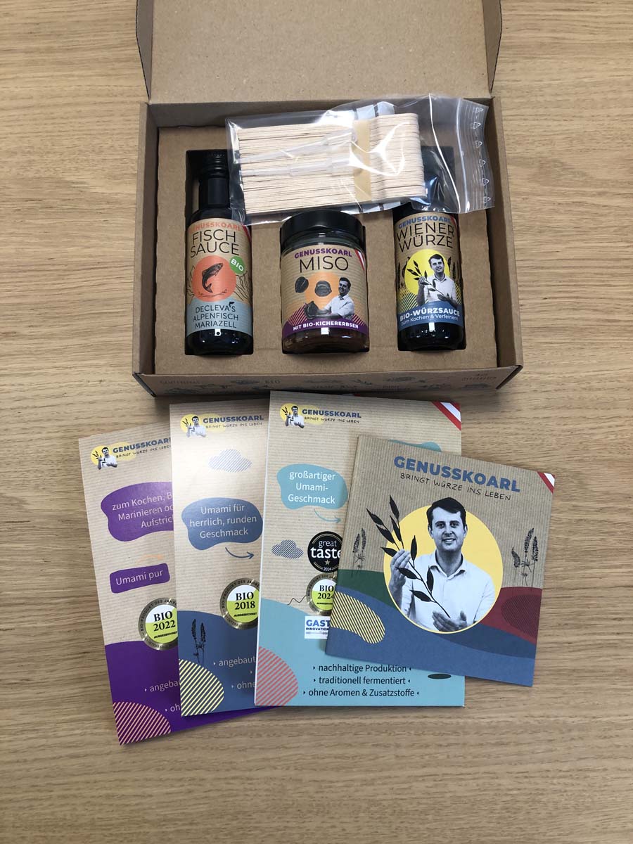 Inhalt der Stillen Verkostungsbox auf einem Tisch ausgelegt: Geschenkbox mit Bio-Fischsauce, Kichererbsen Miso und WienerWürze, Holzspatel, Pipetten und Produktbeschreibungen.