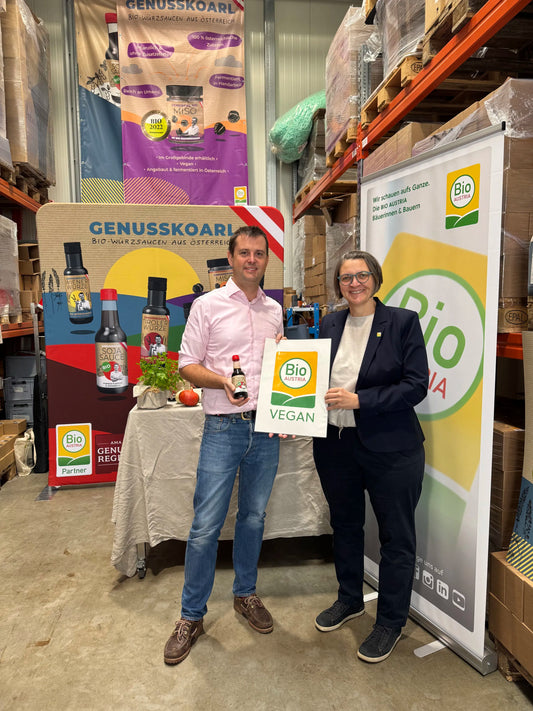 BIO AUSTRIA Obrau Barbara Riegler und Karl Severin Traugott von Genusskoarl präsentieren das BIO AUSTRIA Vegan logo in einer Lagerhalle mit Rollups als Dekor